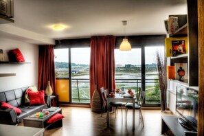 A modern living room with a sofa, dining table, and chairs. A large window offers a view of a river and greenery.