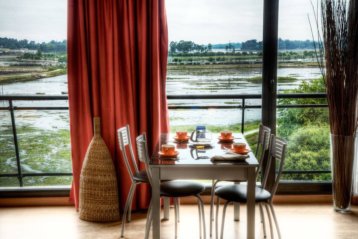 A dining table set with orange dishes, a woven vase, and chairs in a room with a large window overlooking a water body and greenery.