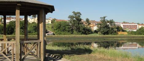 Un quiosco de madera junto a un lago tranquilo, con vistas claras a edificios residenciales al fondo.