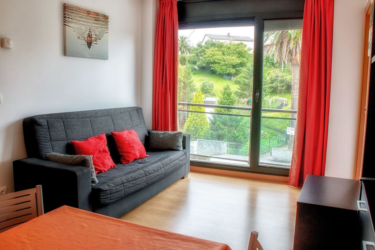 A modern living room with a grey sofa, red cushions, a wooden table, and a large window with red curtains.