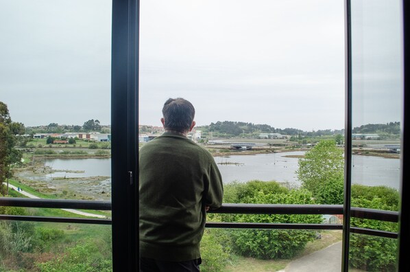 A man standing by a window overlooking a river with a bridge and greenery.