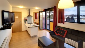 Un appartement moderne comprenant une cuisine, un coin repas et un salon. Il dispose d’une grande fenêtre offrant une vue sur la ville, d’un téléviseur à écran plat et d’un coin salon confortable.