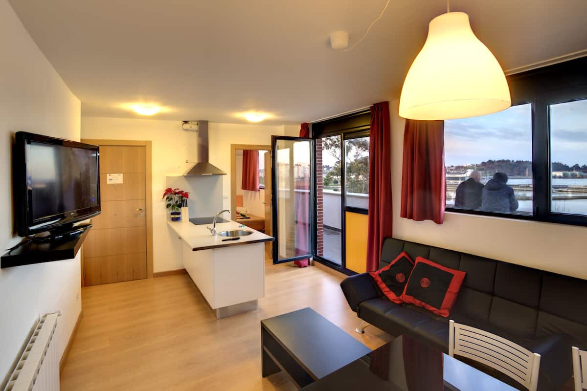 A modern apartment with a kitchen, dining area, and living room. It features a large window with a view of the city, a flat-screen TV, and a comfortable seating area.