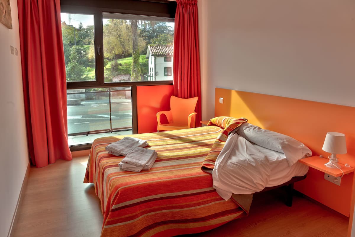A hotel room with two beds, a window with curtains, a chair, and a bedside table with a lamp.