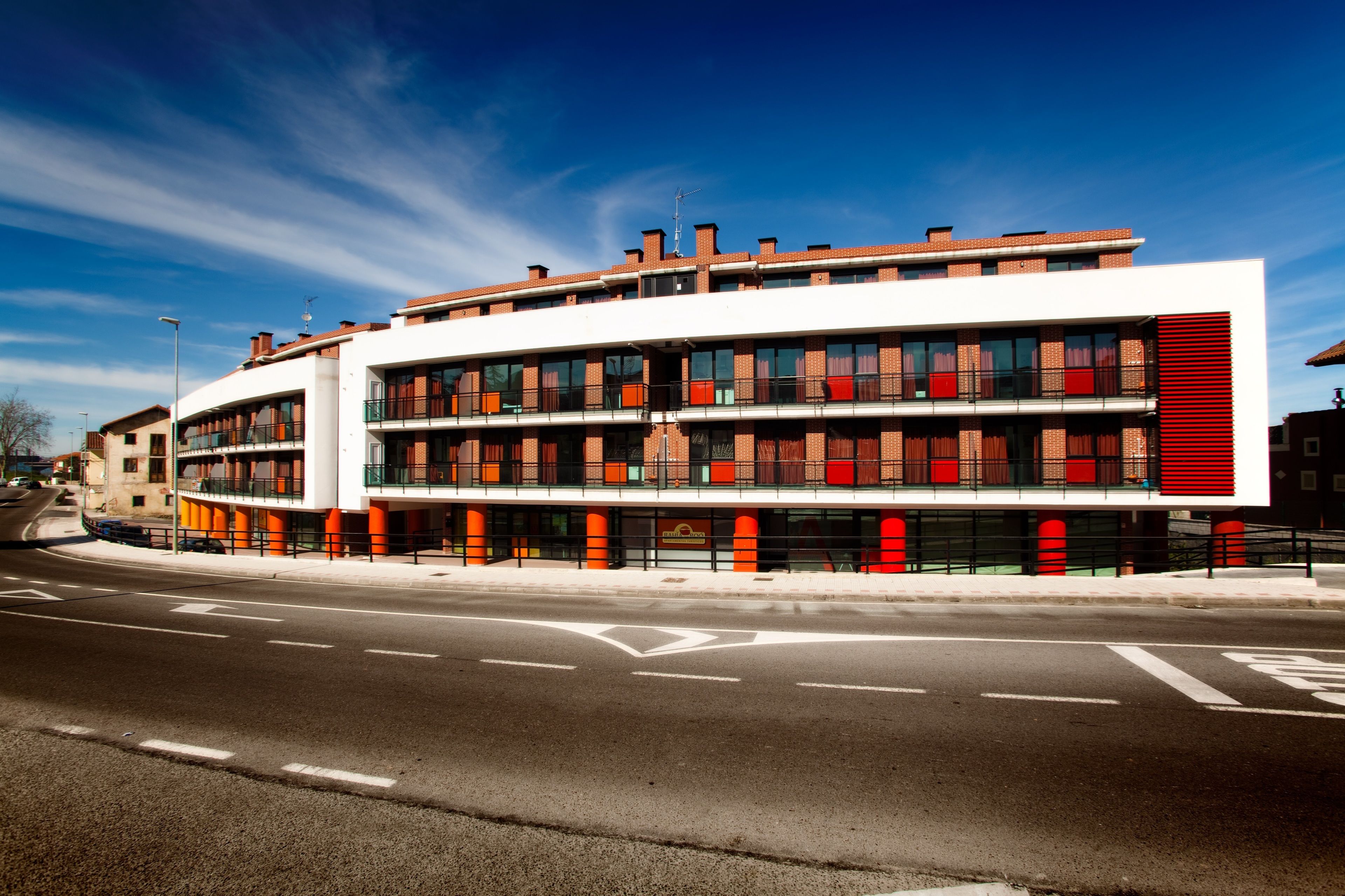Un bâtiment moderne à la façade incurvée, doté de nombreux balcons et d’un mur d’accent rouge bien visible.
