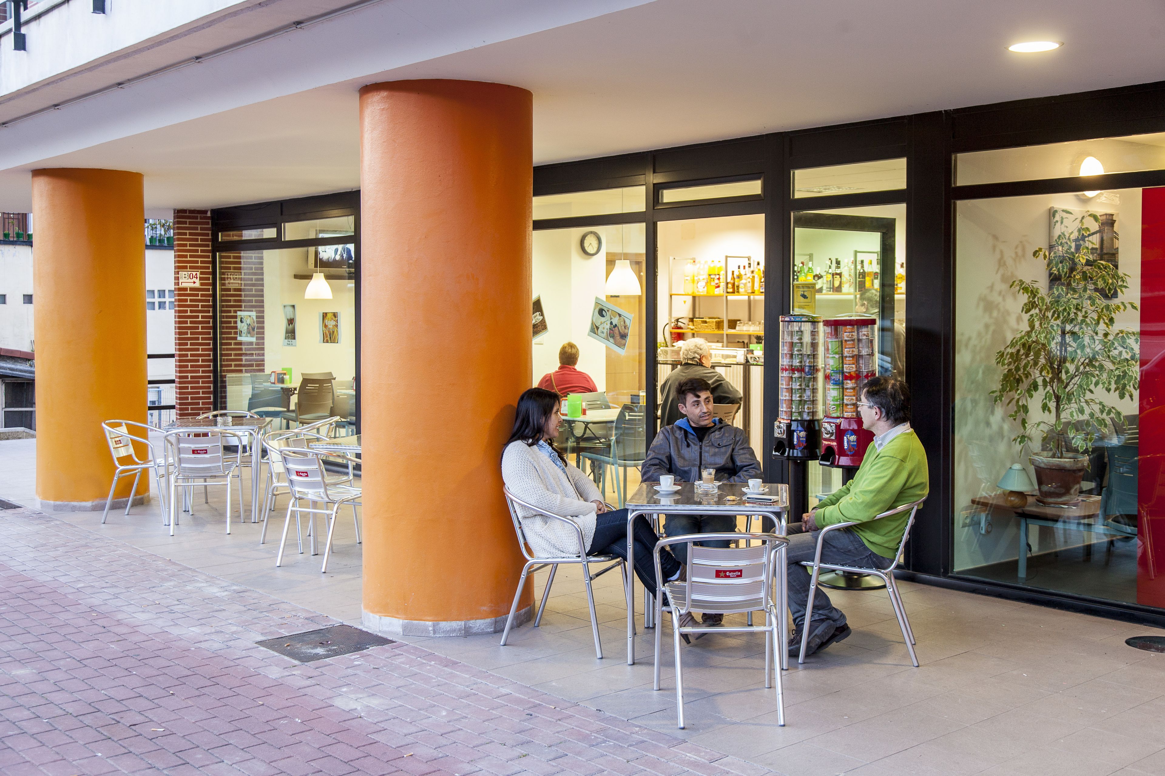 Um café ao ar livre com mesas e cadeiras, uma parede de tijolos e uma fachada de vidro com prateleiras cheias de garrafas.