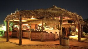 A thatched-roof outdoor bar area with a sandy floor, wooden counters, and a rustic ambiance. - Villaggio Turistico Internazionale La Plaja (Catania)