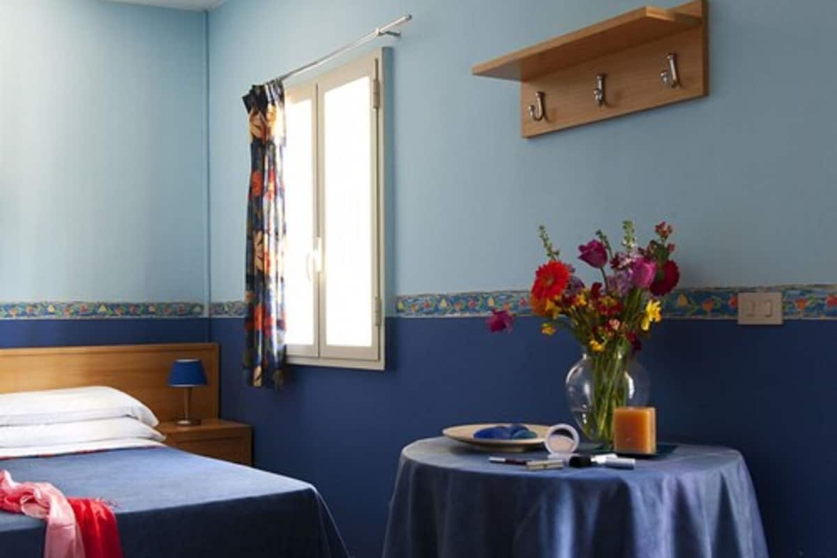 Una habitaci ón con dos camas, una ventana con cortina, una mesita con un florero y un estante con ganchos.