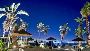 A beachside lounge area with sun loungers and umbrellas, palm trees, and a view of the city lights at night. - Villaggio Turistico Internazionale La Plaja (Catania)
