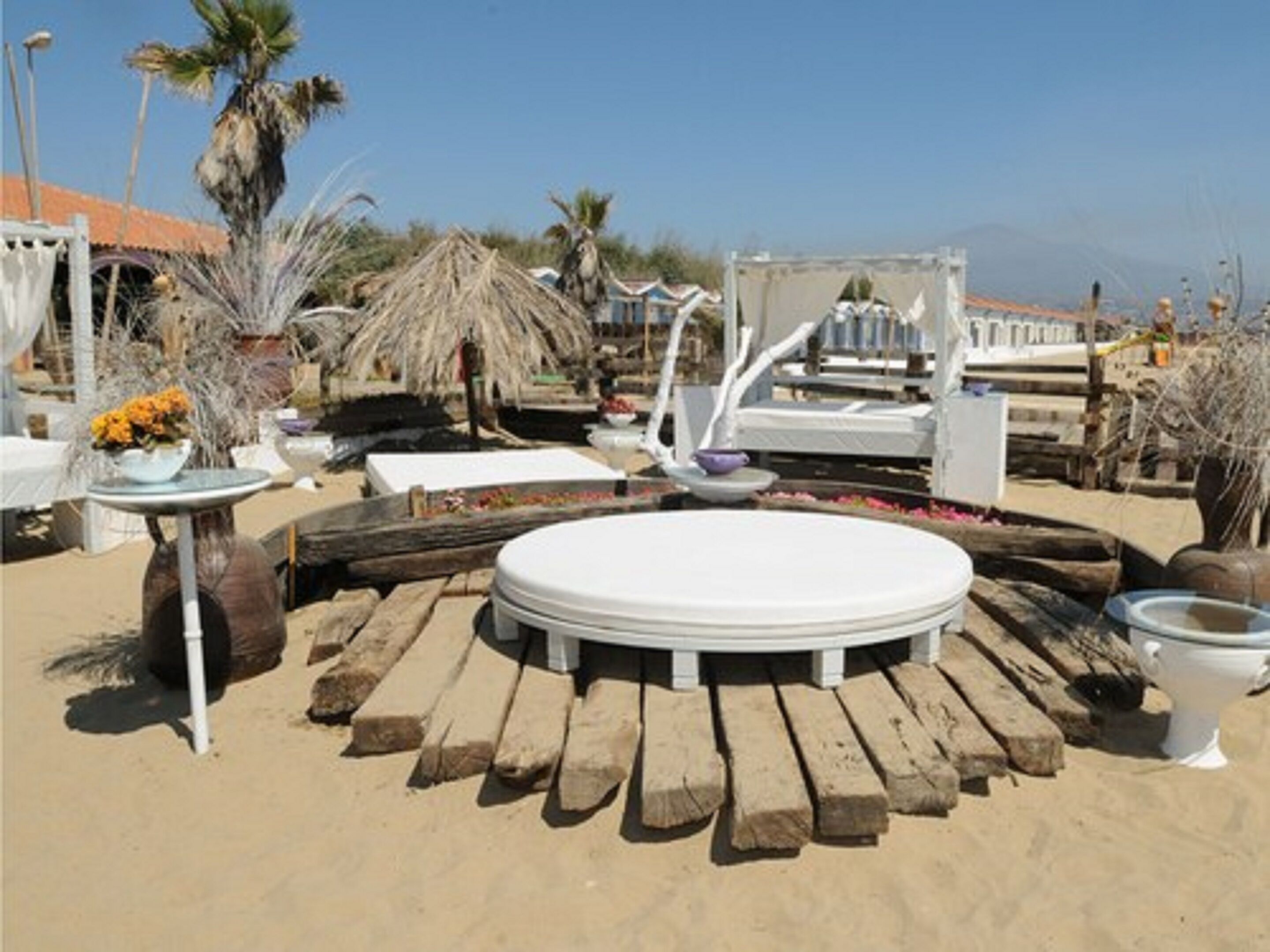 Een terras aan het strand met een ronde houten tafel, witte stoelen en een wit toilet.