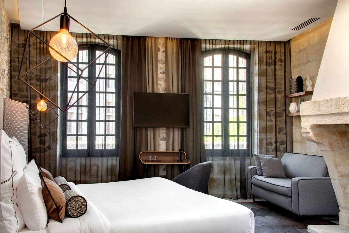 A modern hotel room with a large bed, a sofa, a TV, and large windows with curtains.