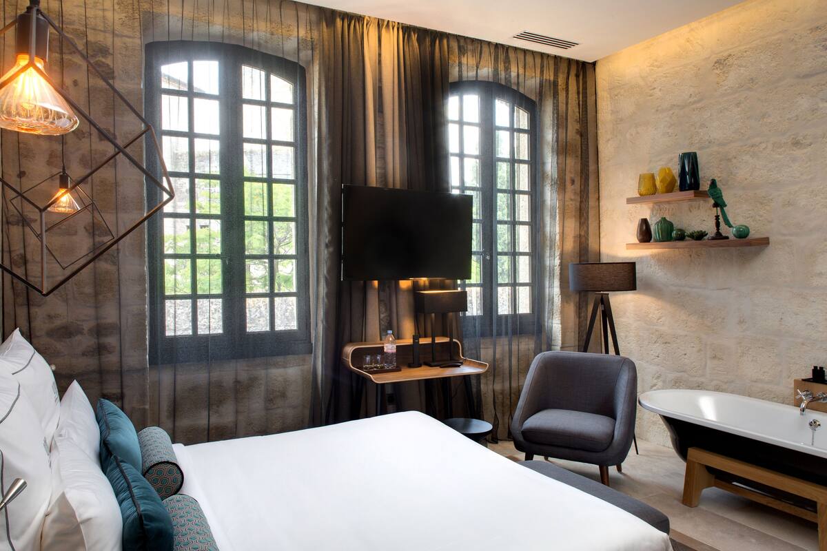 A modern hotel room with a large bed, a desk, a flat-screen TV, a chair, a bathtub, and a shelf with decorative items.