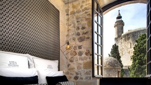 A bedroom with a stone wall, a bed with patterned pillows, and a view of a historic building through an open window. - Boutique Hôtel des Remparts (Aigues-Mortes)
