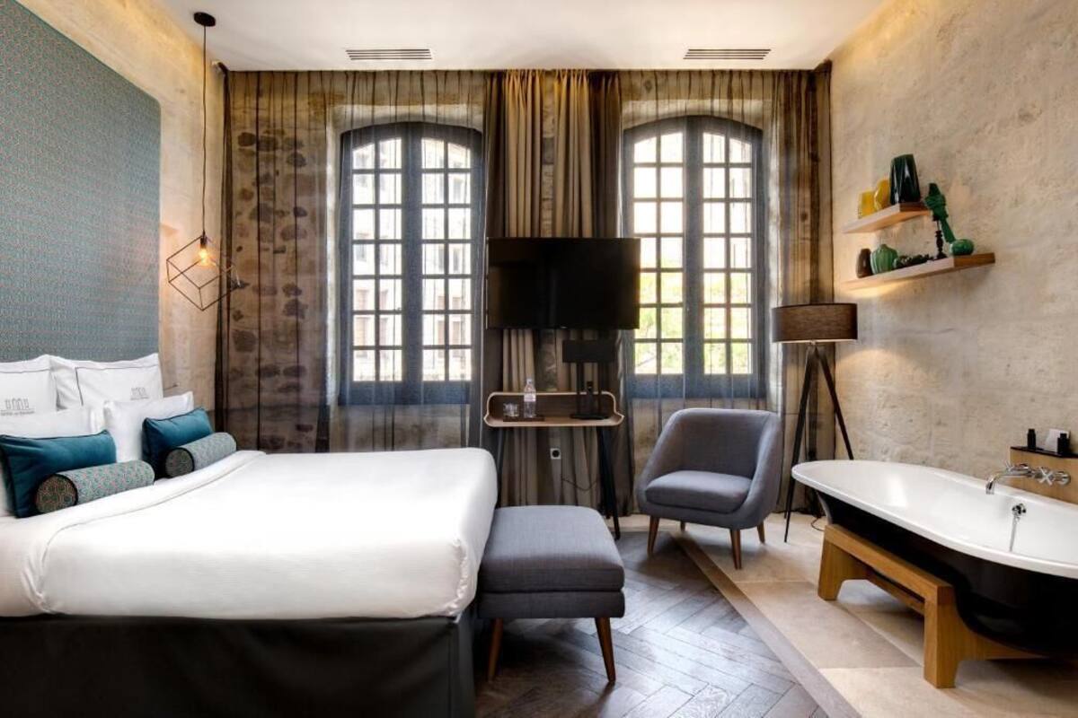 A hotel room with a bed, a chair, a desk, a television, a shelf with toiletries, and a bathtub.