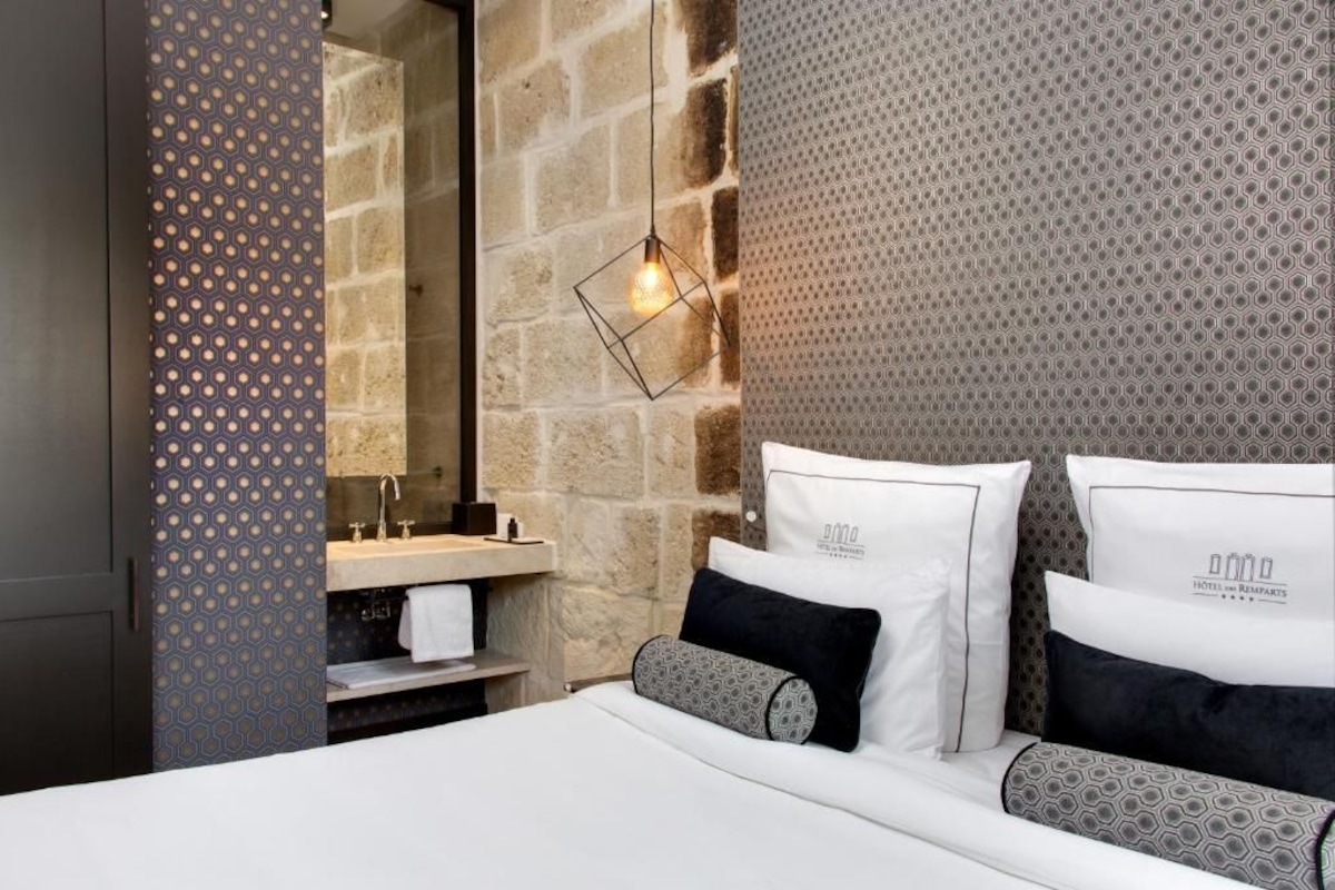 Quarto de hotel com parede de pedra, cama com almofadas estampadas, luminária pendente e banheiro com espelho e pia.