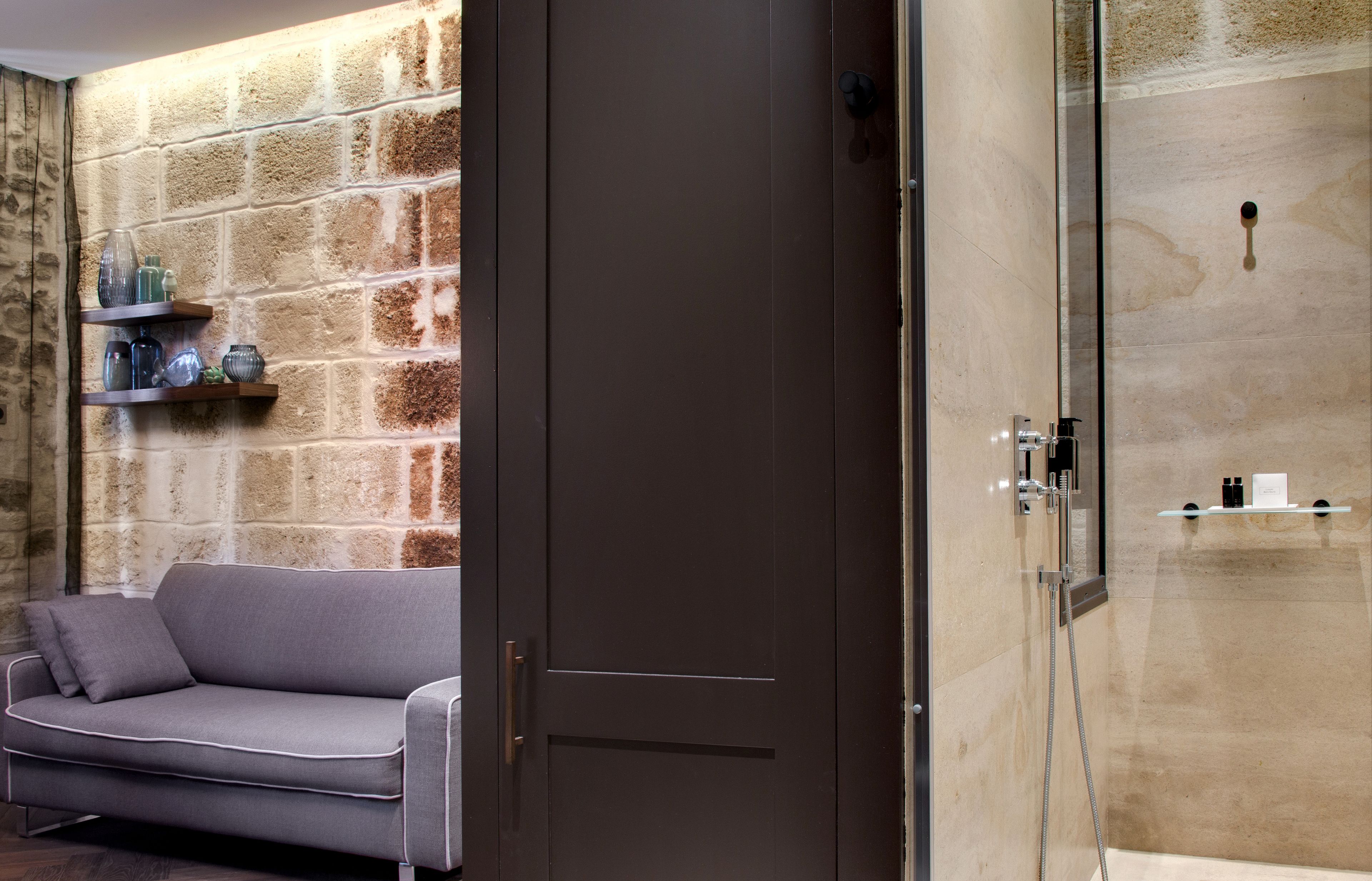 A modern bathroom with a glass shower enclosure, a beige tiled wall, and a grey sofa with a shelf above it.