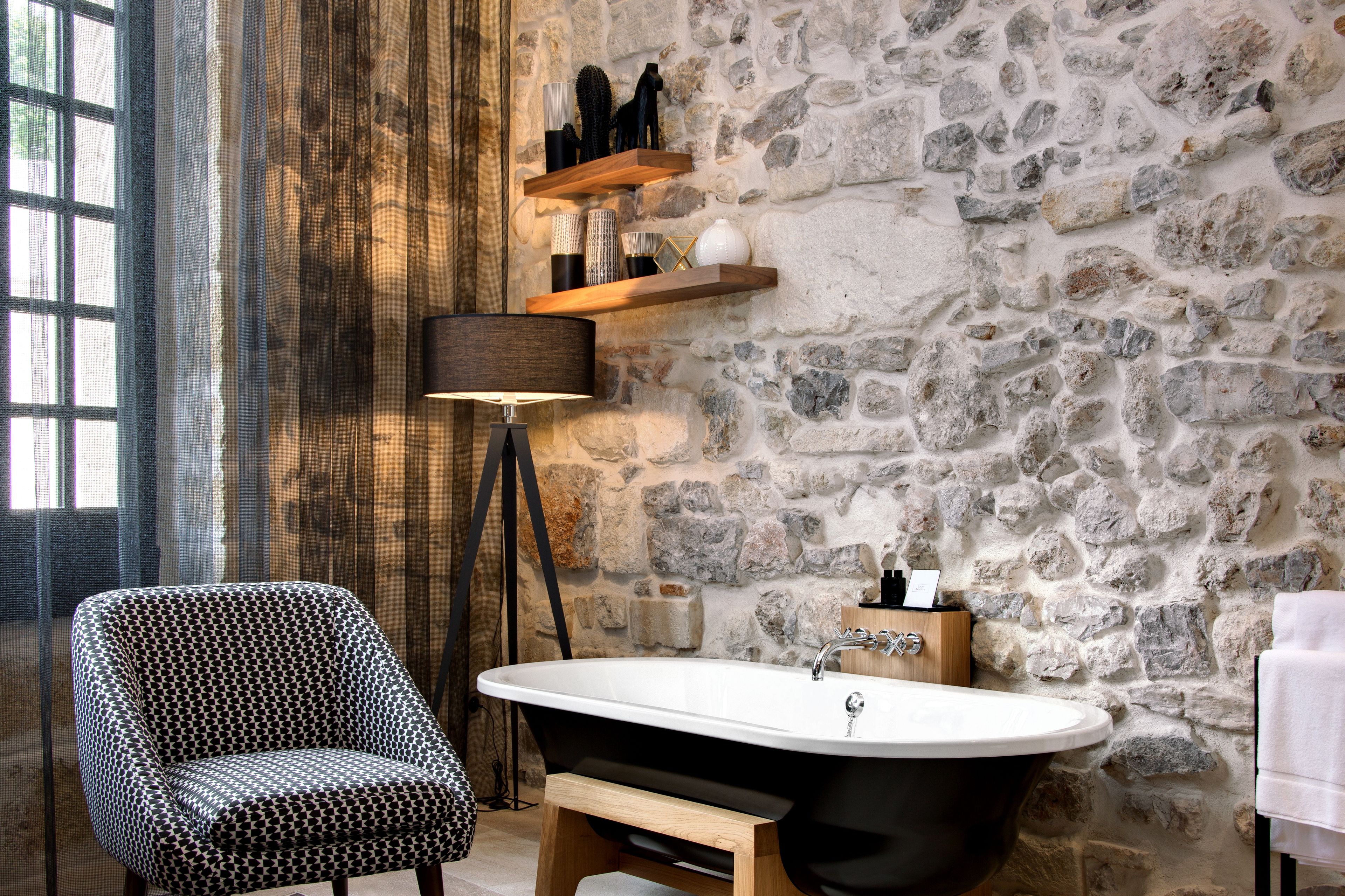 Un baño con pared de piedra, una bañera independiente, un sillón estampado y una lámpara de pie.