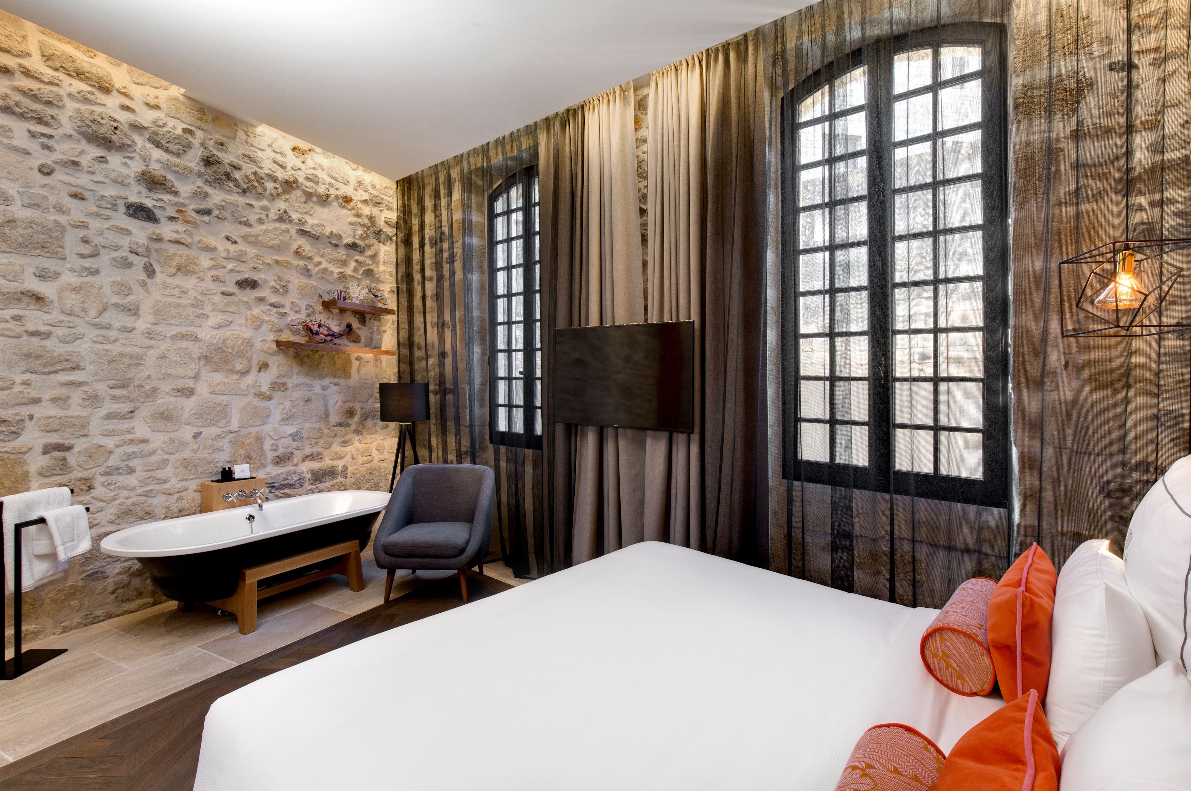 A room with a stone wall, a bathtub, a bed with white bedding, and a large window with curtains.