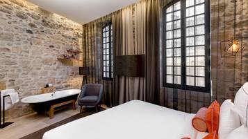 A room with a stone wall, a bathtub, a bed with white bedding, and a large window with curtains.