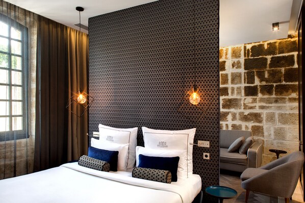 A modern hotel room with a large bed, two bedside tables, a sofa, and a stone accent wall. - Boutique Hôtel des Remparts (Aigues-Mortes)