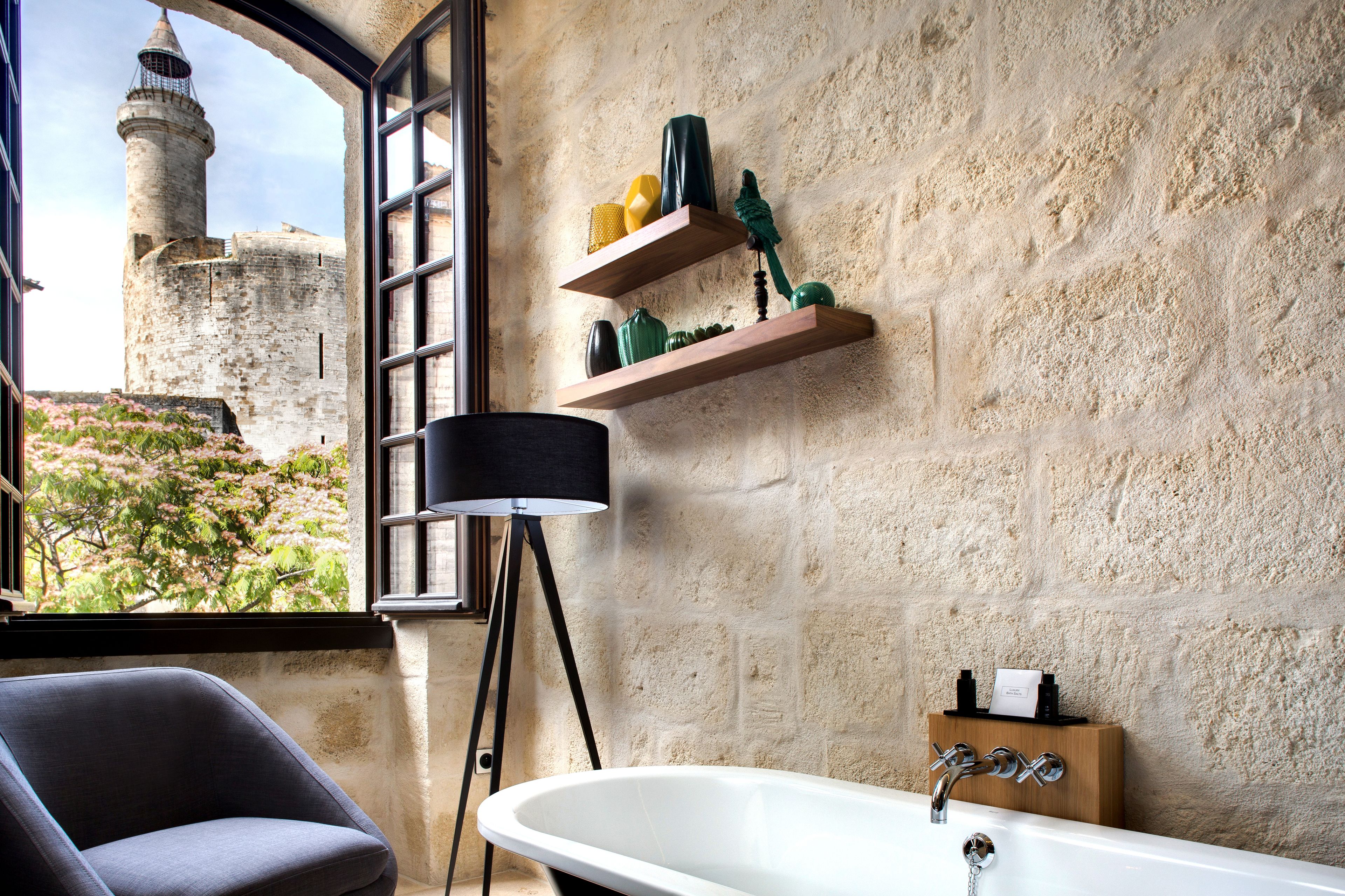 A bathroom with a large window, a bathtub, a chair, and wooden shelves with toiletries.