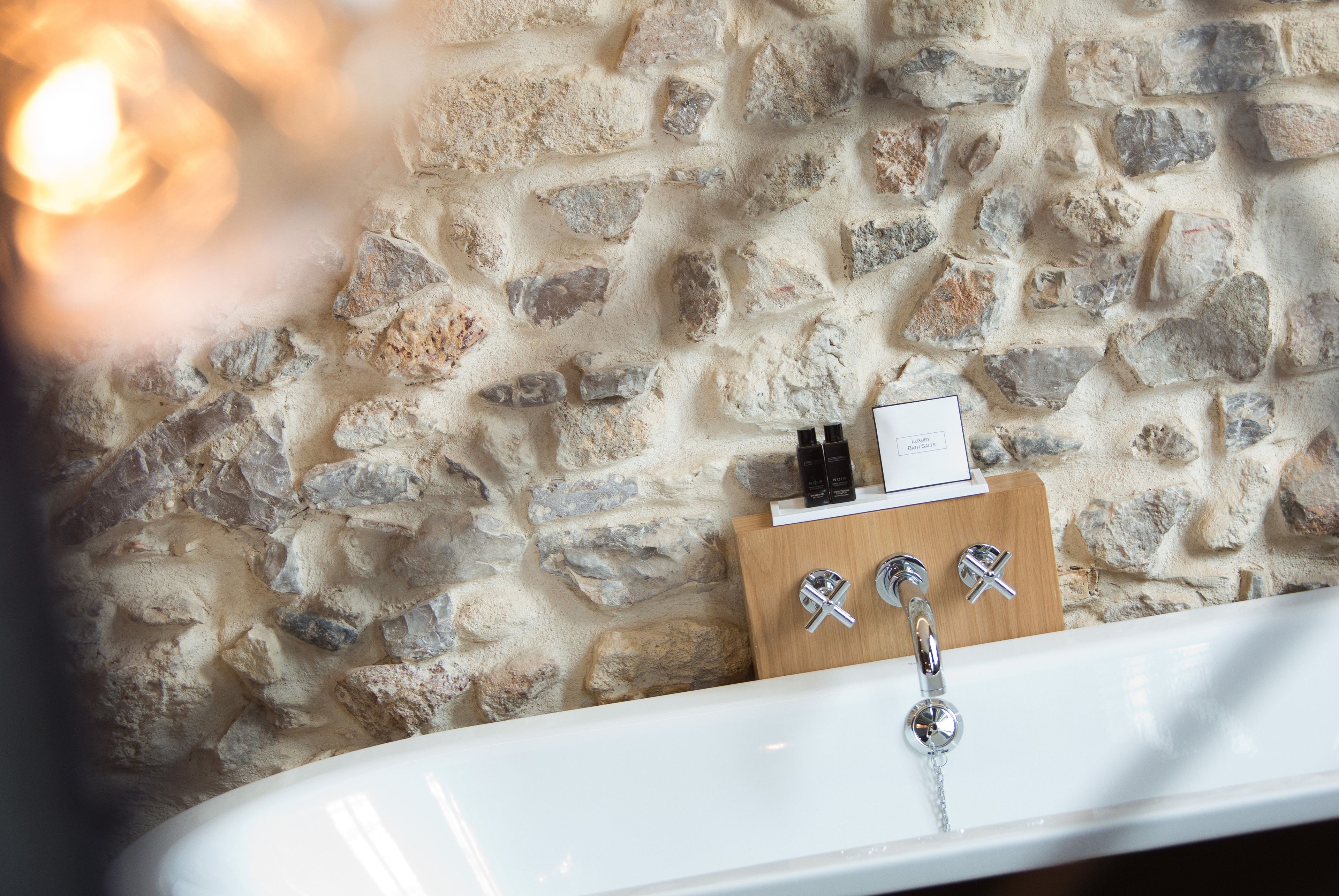 Une salle de bain avec un mur en pierre, une baignoire blanche et une étagère en bois où sont rangés les articles de toilette.