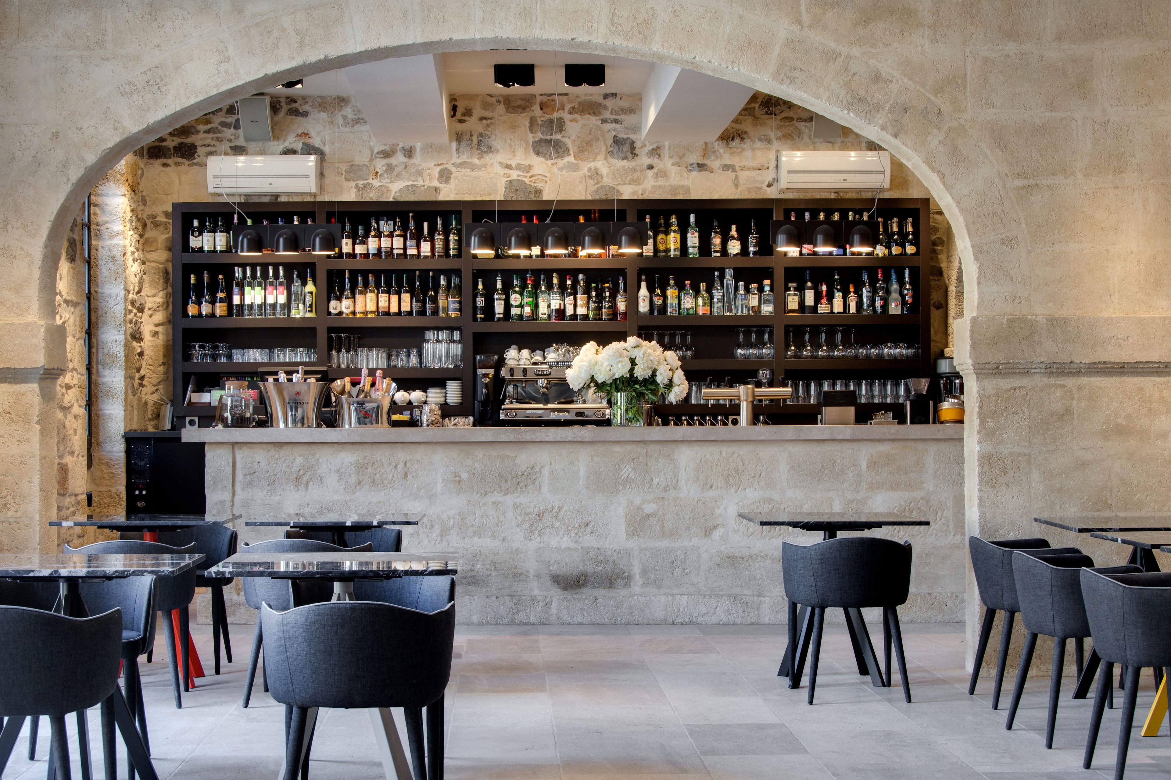A bar area with a stone wall, shelves of liquor bottles, and a floral centerpiece. Tables and chairs are arranged in front.