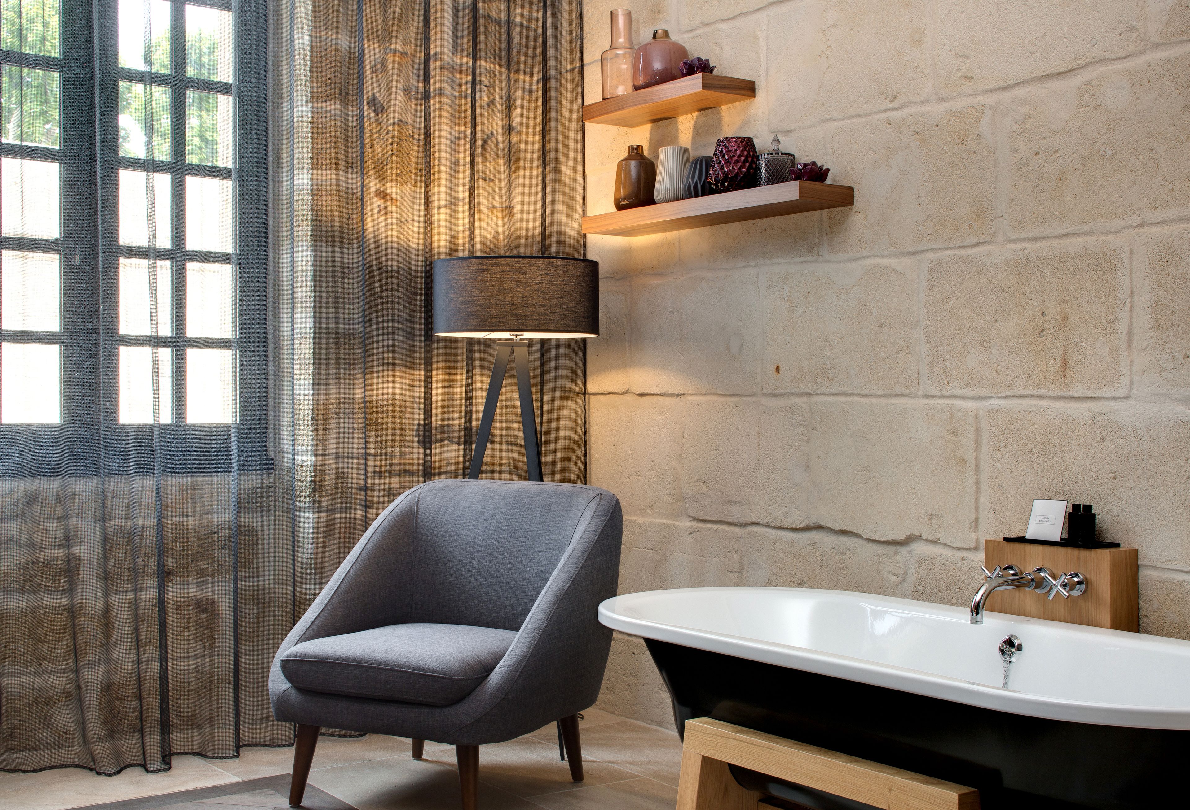 Un baño con bañera, una silla, una lámpara y repisas con objetos decorativos.