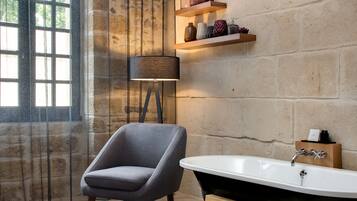Un baño con bañera, una silla, una lámpara y repisas con objetos decorativos.