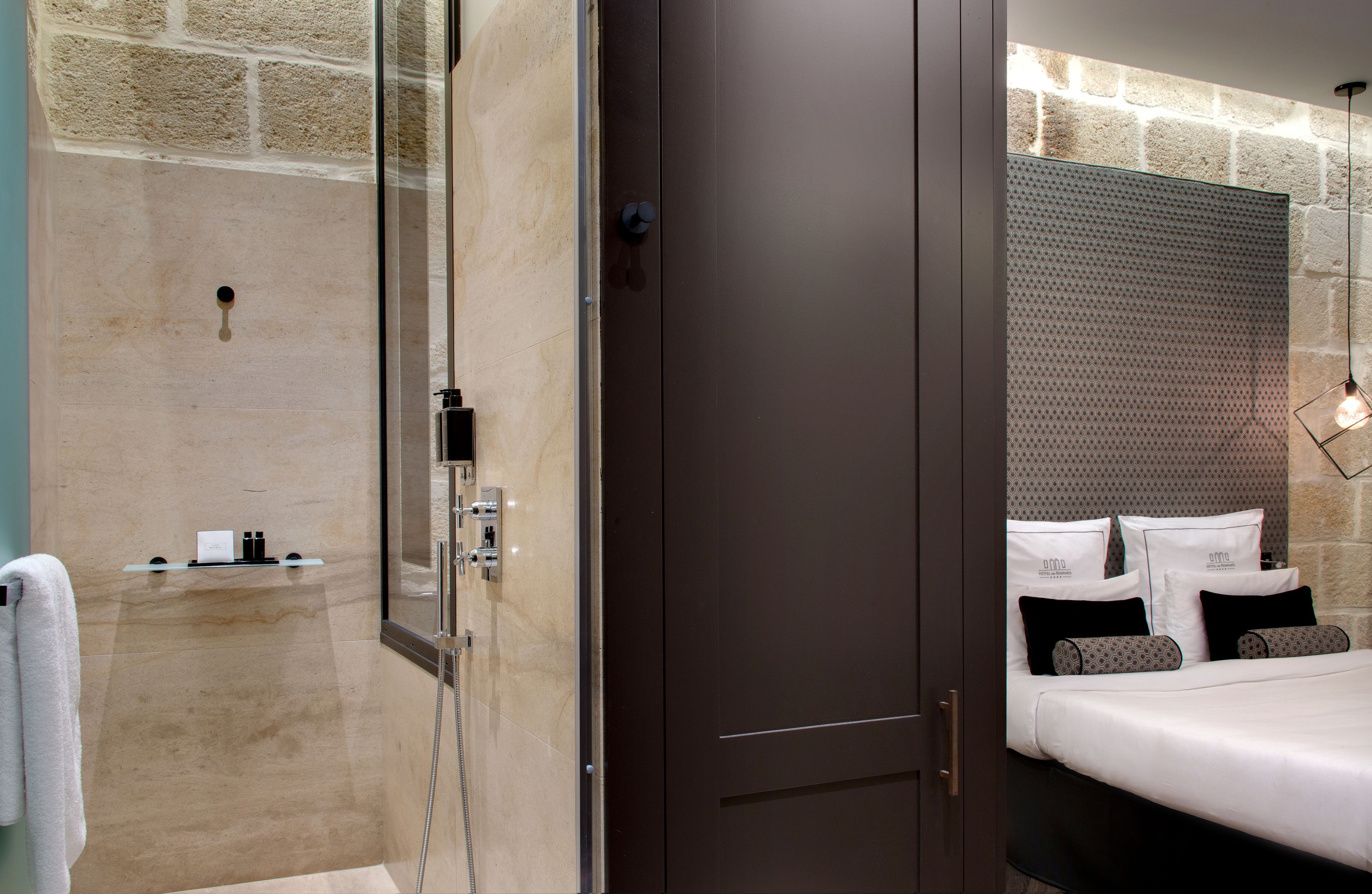 A modern hotel room with a glass-enclosed shower, a dark wooden door, a bed with white bedding and patterned pillows, and a stone wall.