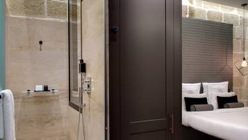 A modern hotel room with a glass-enclosed shower, a dark wooden door, a bed with white bedding and patterned pillows, and a stone wall.