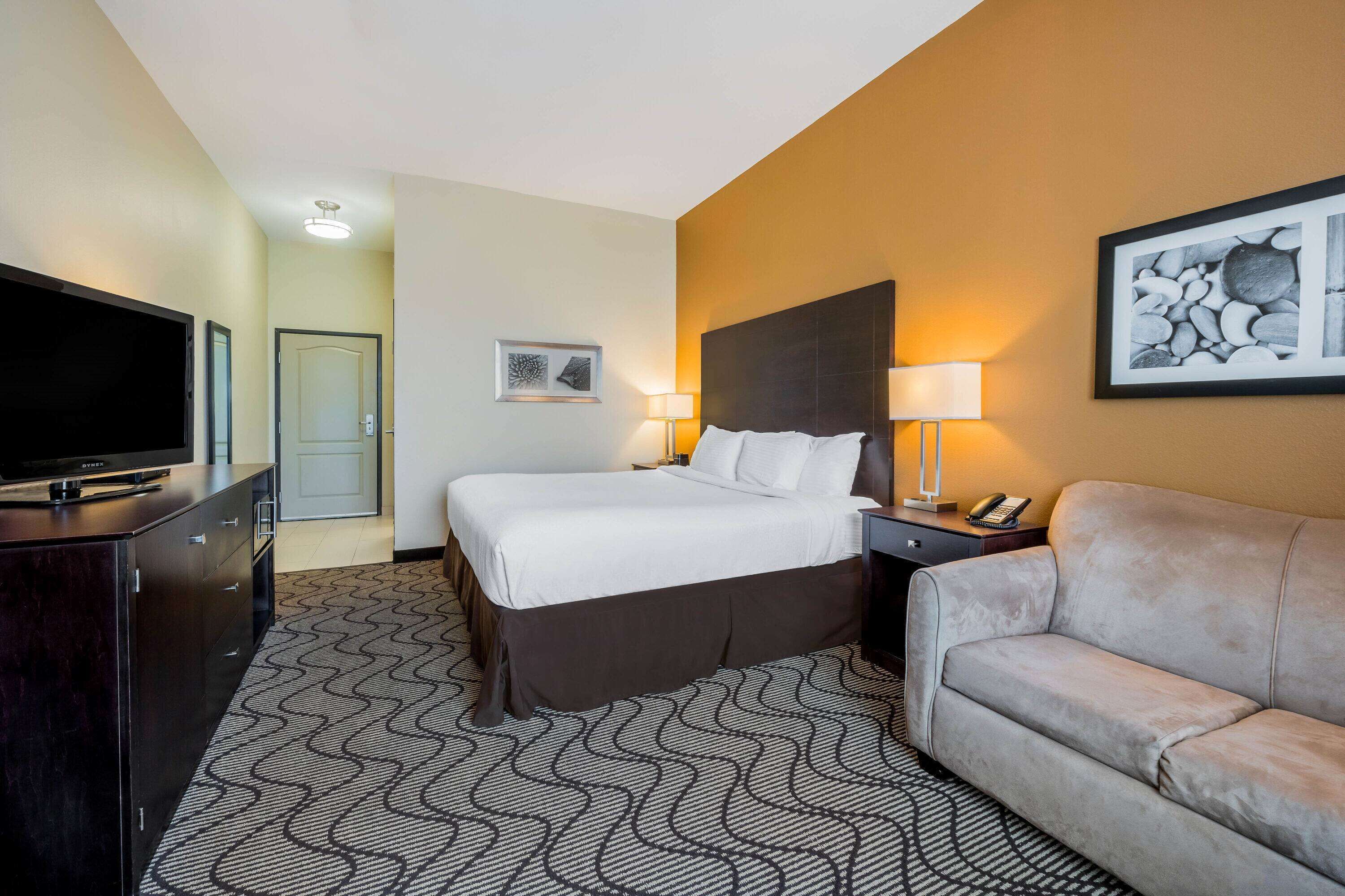 A hotel room with a bed, a TV, a sofa, a desk with a lamp, and a picture on the wall.