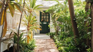 A brick pathway leading to a green door, surrounded by lush tropical plants and a white fence.