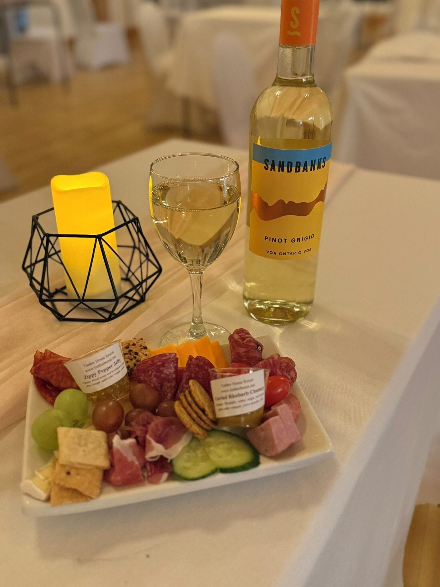 Una mesa con una copa de vino blanco, una botella de Pinot Grigio y una tabla de embutidos, quesos y frutas.