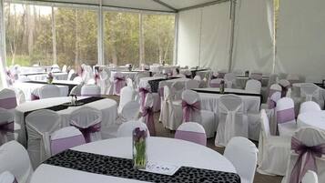 Un espace événementiel aménagé sous une tente, avec des tables rondes, des chaises blanches ornées de rubans violets et un lustre.