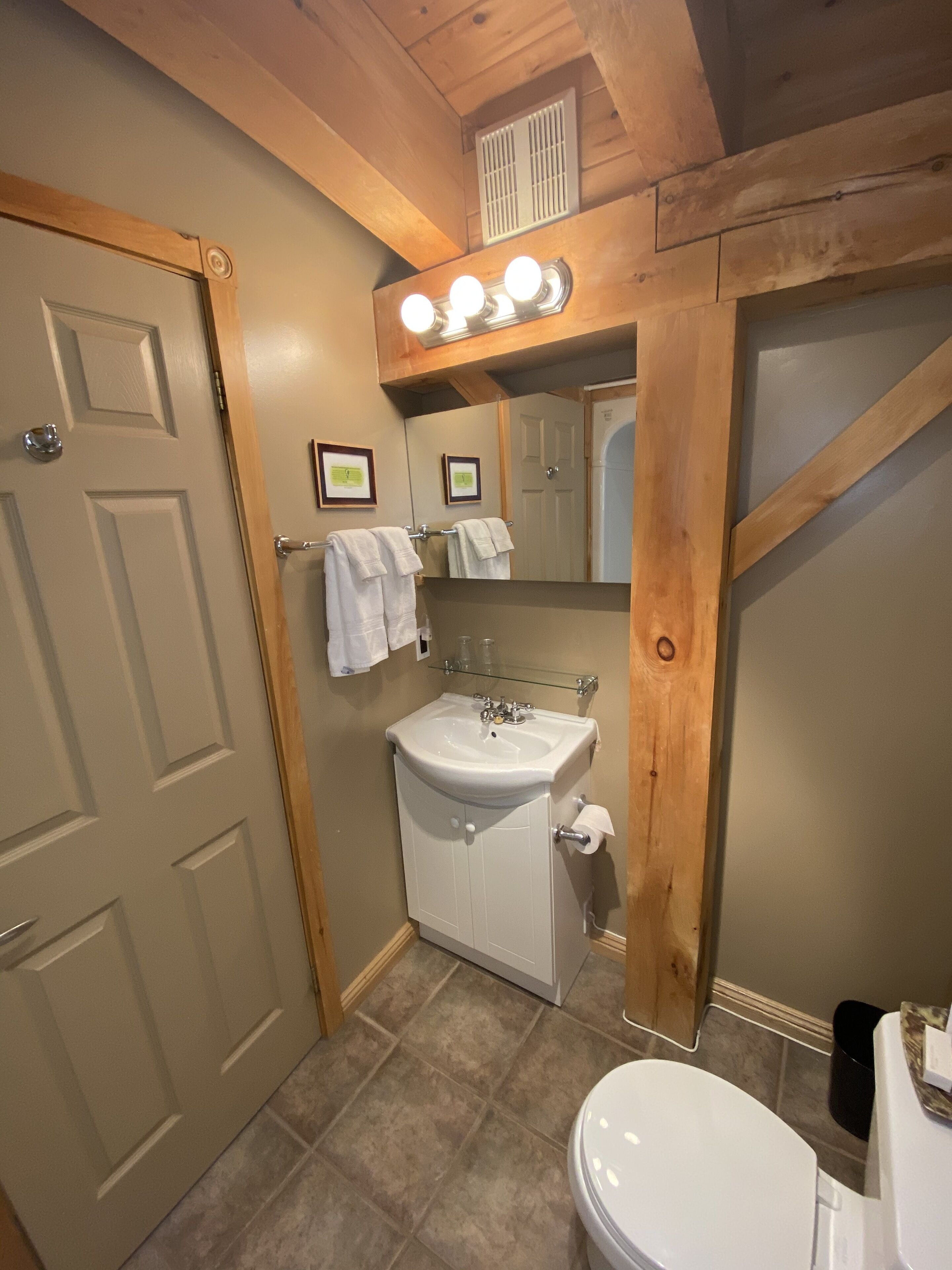 A bathroom with a white sink, a mirror, and a toilet. There are towels hanging on the wall and a window with blinds.