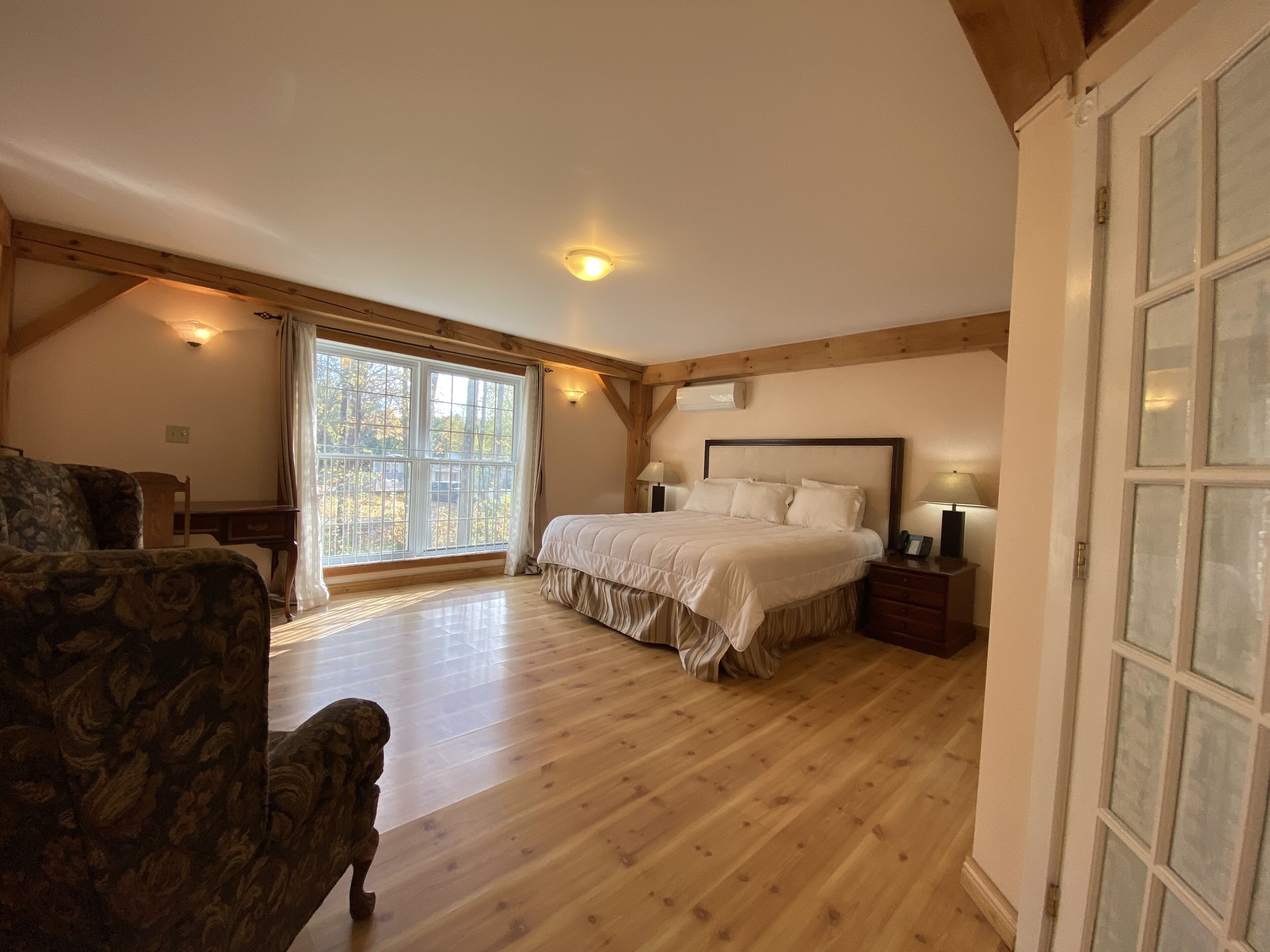 A bedroom with a large bed, wooden beams, a window with curtains, and a chair.