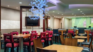 Een restaurantruimte in een hotel met meerdere tafels en stoelen, een aan de muur bevestigde flatscreen televisie en een plafond met een decoratieve verlichtingsarmatuur.