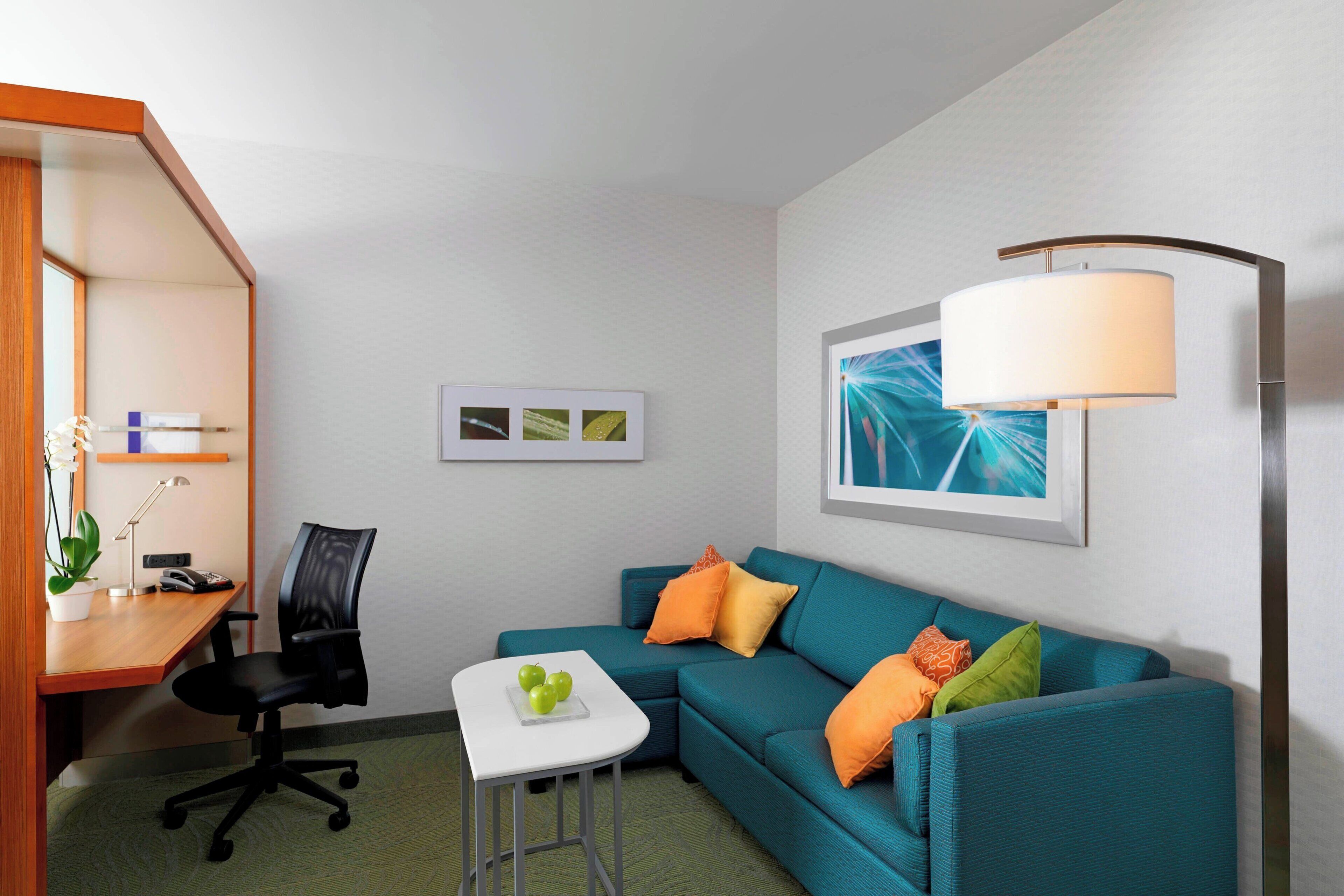 A modern hotel room with a teal sofa, a small white table with a fruit bowl, a black office chair, a desk with a lamp, and a wall-mounted lamp.