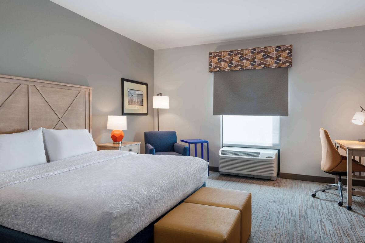 A hotel room with a large bed, a desk, a chair, a small table, a lamp, and a window with a view.