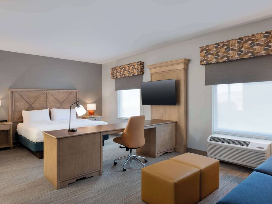 A hotel room with a bed, desk, chair, TV, air conditioning unit, and large windows.