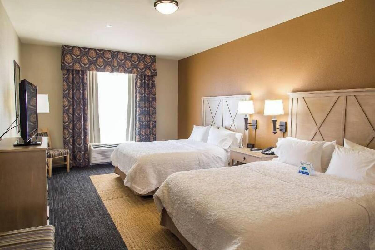 A hotel room with two beds, a desk, a television, and a window with patterned curtains.