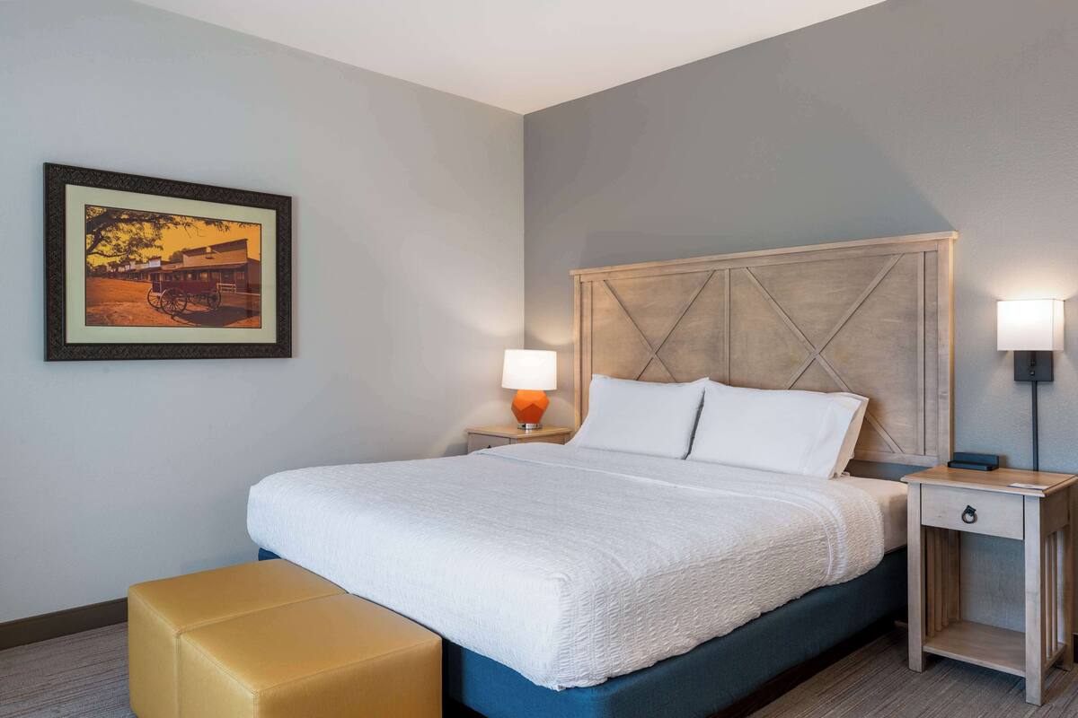 A neatly made bed with a headboard, two bedside tables with lamps, a framed picture on the wall, and a bench.