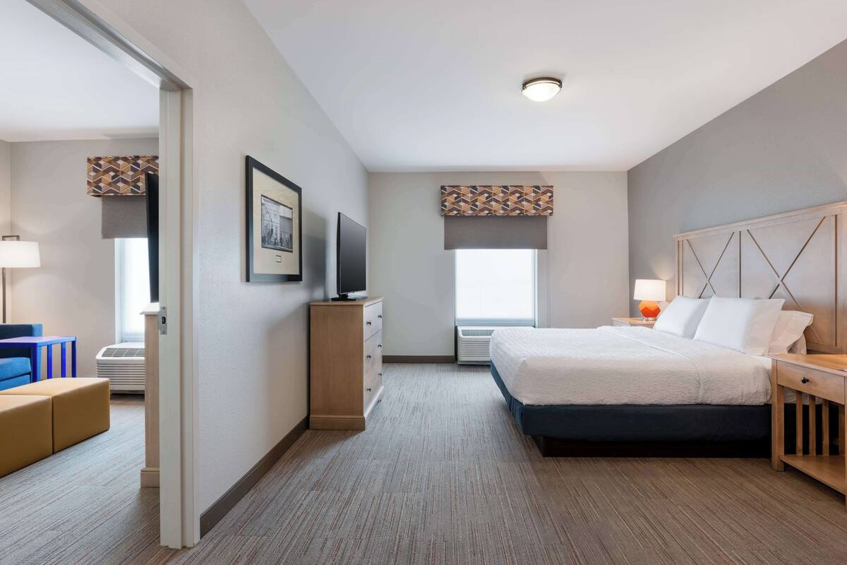 A hotel room with a bed, bedside table, lamp, TV, and a view of the outside.