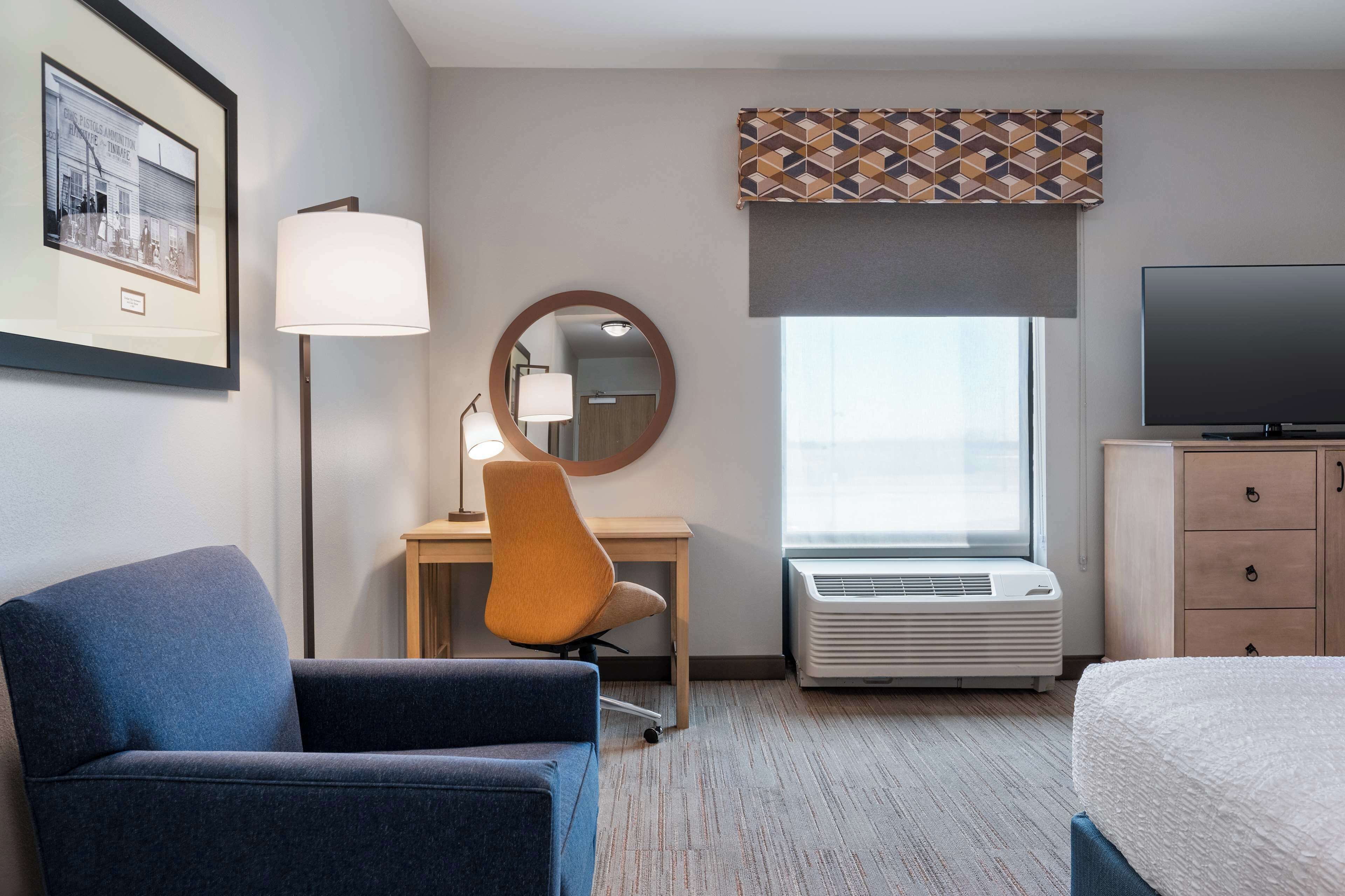 A hotel room with a bed, a desk with a chair, a television, a framed picture, a lamp, and an air conditioner.