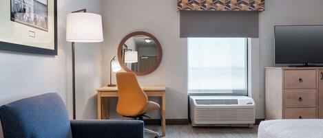 Una habitación de hotel con una cama, un escritorio con una silla, un televisor, un cuadro enmarcado, una lámpara y un aire acondicionado.