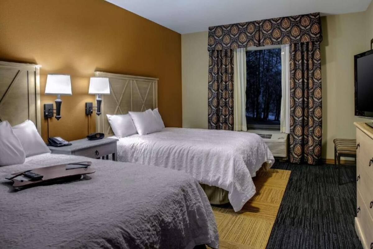 A hotel room with two beds, bedside lamps, a TV, a window with patterned curtains, and a small desk with a phone.