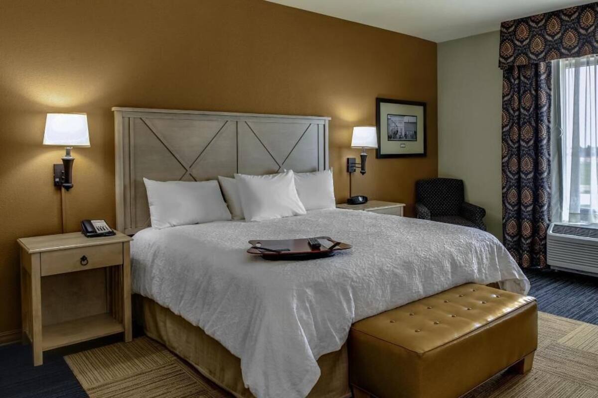 A hotel room with a large bed, two bedside tables, a chair, a window with curtains, and a wall-mounted picture.
