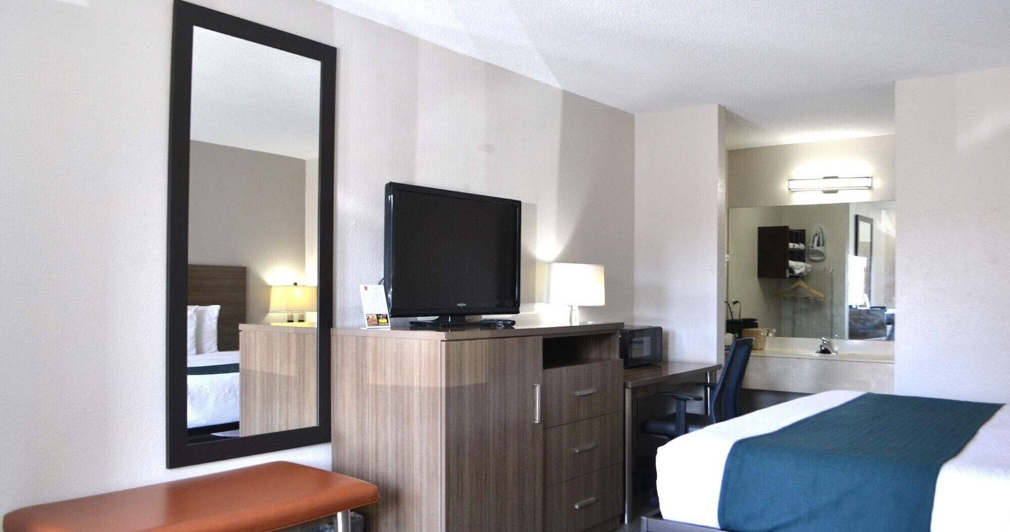 Une chambre d’hôtel avec un lit, un bureau avec un ordinateur, une télévision, un miroir et une salle de bain visible à travers une porte ouverte.