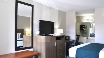 Une chambre d’hôtel avec un lit, un bureau avec un ordinateur, une télévision, un miroir et une salle de bain visible à travers une porte ouverte.
